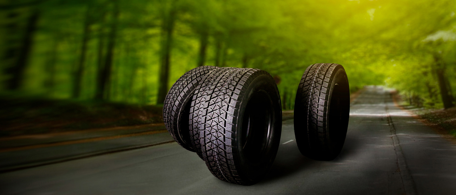 Fire sorte bildæk står oprejst på en tom vej i en grøn skov, hvor solstråler smukt filtrerer gennem trætoppene.