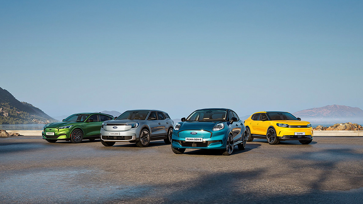 Fire farverige biler, grøn, sølv, blå og gul, er parkeret side om side udendørs med bjerge og en blå himmel i baggrunden.