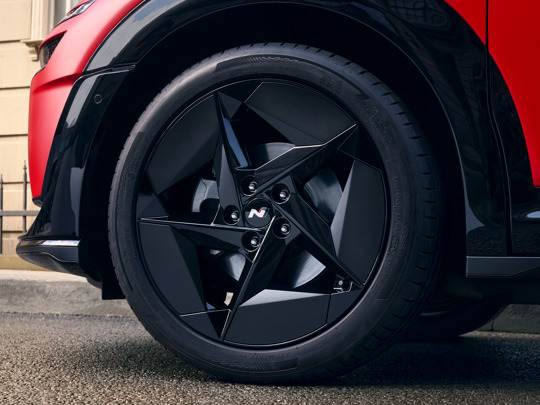 Nærbillede af en sort alufælg med et geometrisk egerdesign på en rød IONIQ 3, der holder parkeret på en gade.