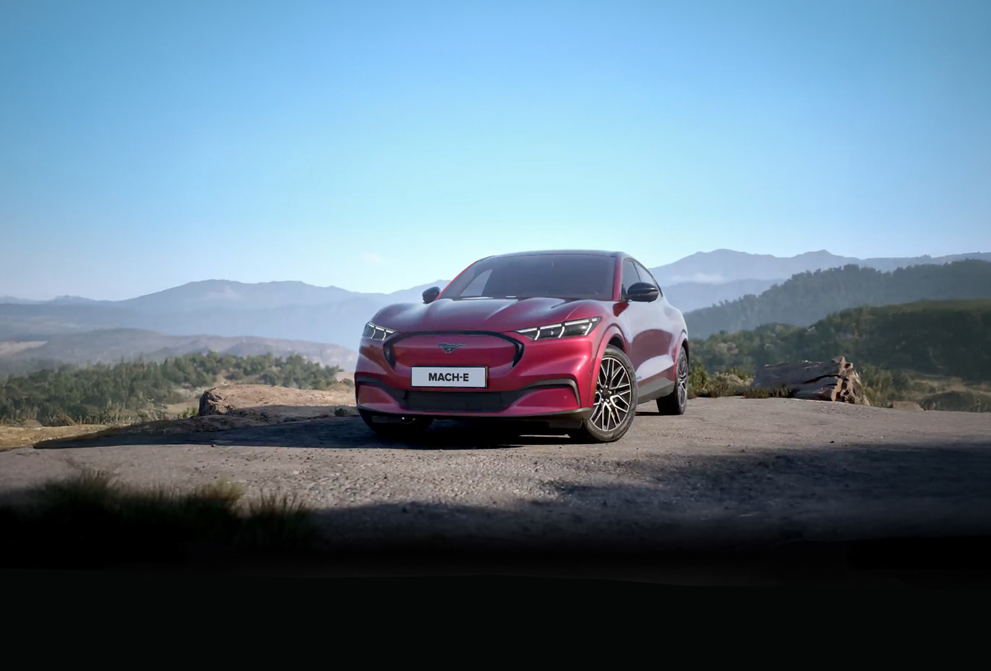 En rød Ford Mustang Mach-E er parkeret på et asfalteret område med bjerge og træer i baggrunden under en klar blå himmel.