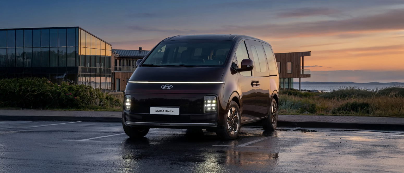 En Hyundai Staria Electric varebil er parkeret på en udendørs parkeringsplads ved solnedgang med moderne bygninger og kystlandskab i baggrunden.