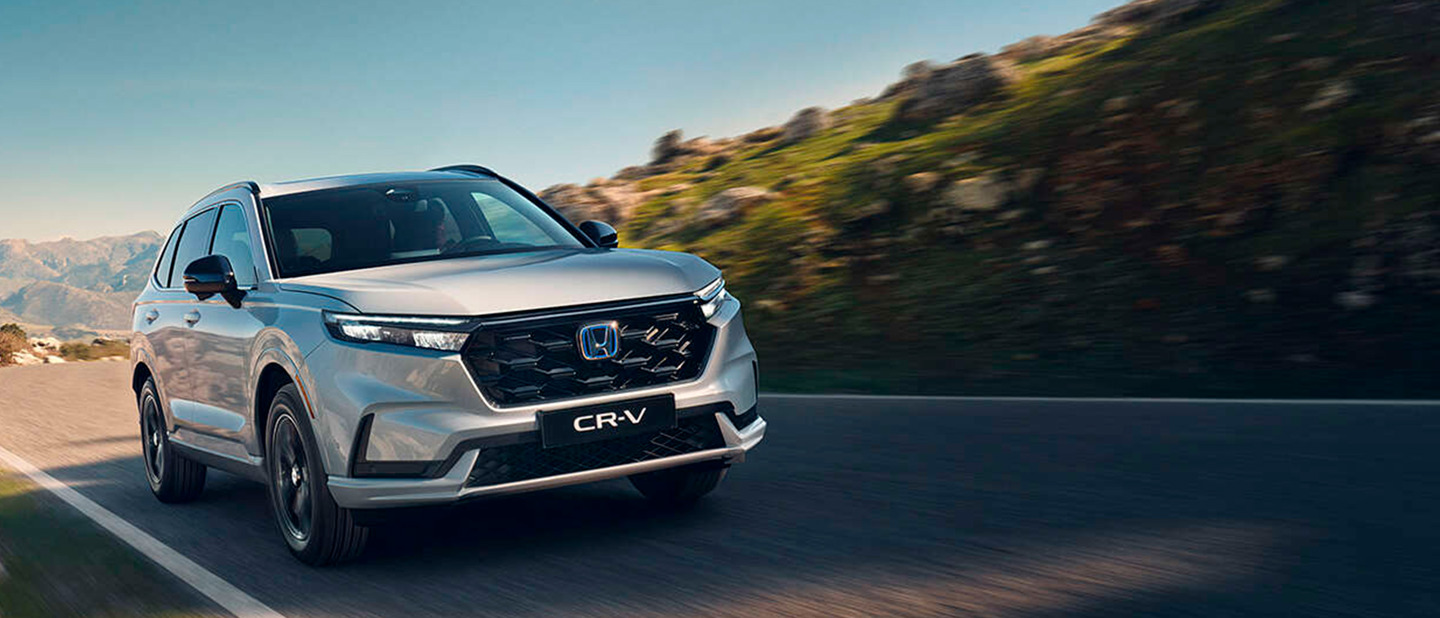 En sølvfarvet Honda CR-V kører på en snoet vej med stenede bakker og klar himmel i baggrunden.