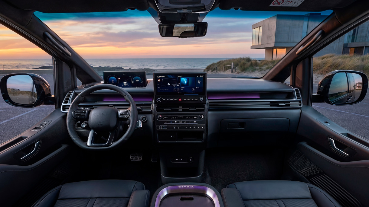 Moderne bilinteriør med digitale skærme, lædersæder og havudsigt gennem forruden ved solnedgang; moderne hus synligt udenfor.