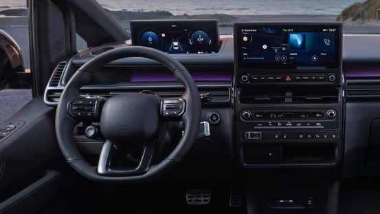 Moderne bilinteriør med digitalt instrumentbræt, touch-skærme, rat og kontrolknapper. Udsigt til havet gennem forruden.