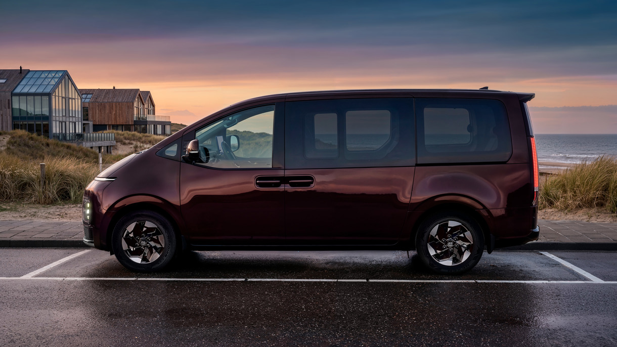 En mørk minivan parkeret i vejkanten nær moderne huse og klitter ved solnedgang, med stranden og havet i baggrunden.