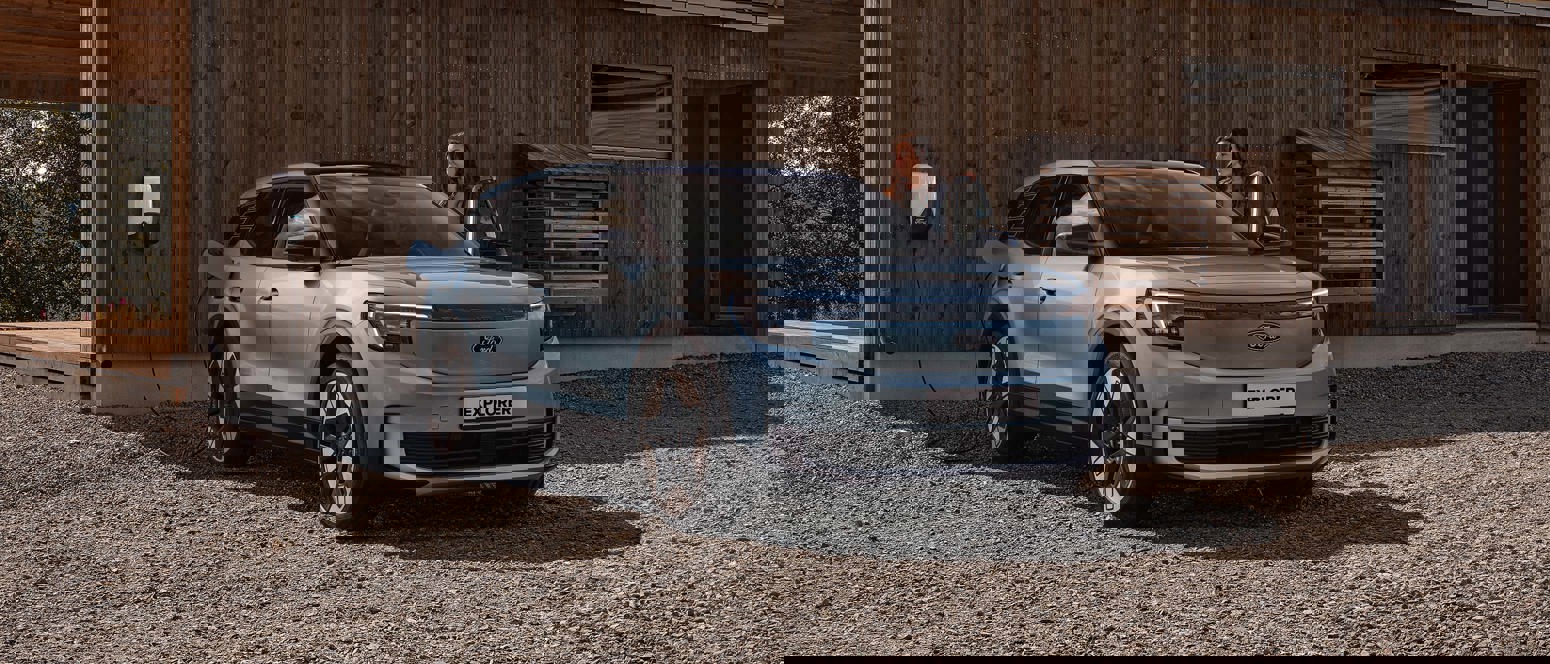 En kvinde står ved siden af en blå Ford Explorer el-SUV, der er parkeret ved en træbygning og oplader ved en udendørs station.