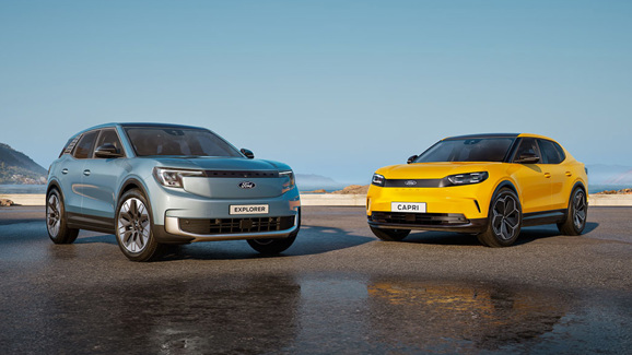 To elektriske SUV'er fra Ford, Explorer og Capri, er parkeret side om side udendørs på en klar dag med bjerge i baggrunden.