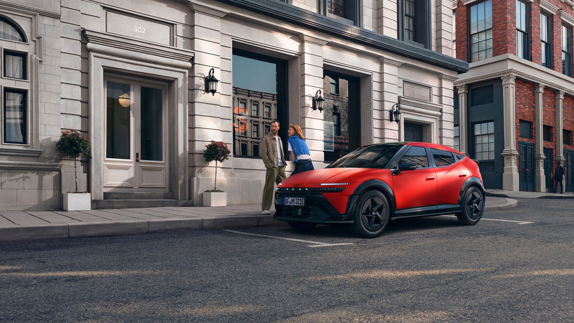 En rød elektrisk IONIQ 3 er parkeret på en gade i byen i nærheden af to personer, der står uden for en bygning med store vinduer.