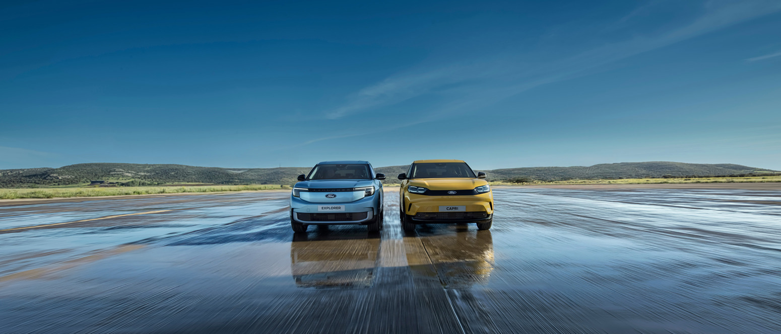 To moderne SUV'er, en sølvfarvet og en gul, parkeret side om side på en våd, reflekterende overflade med bakker og blå himmel i baggrunden.