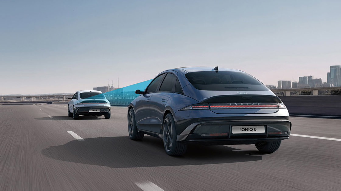 To moderne biler kører på en motorvej; den ene viser en blå sensorstråle, hvilket tyder på avanceret førerassistanceteknologi.