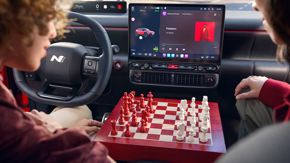 To personer spiller skak på et rødt og hvidt bræt inde i en IONIQ 3, mens der afspilles musik på bilens touchscreen.